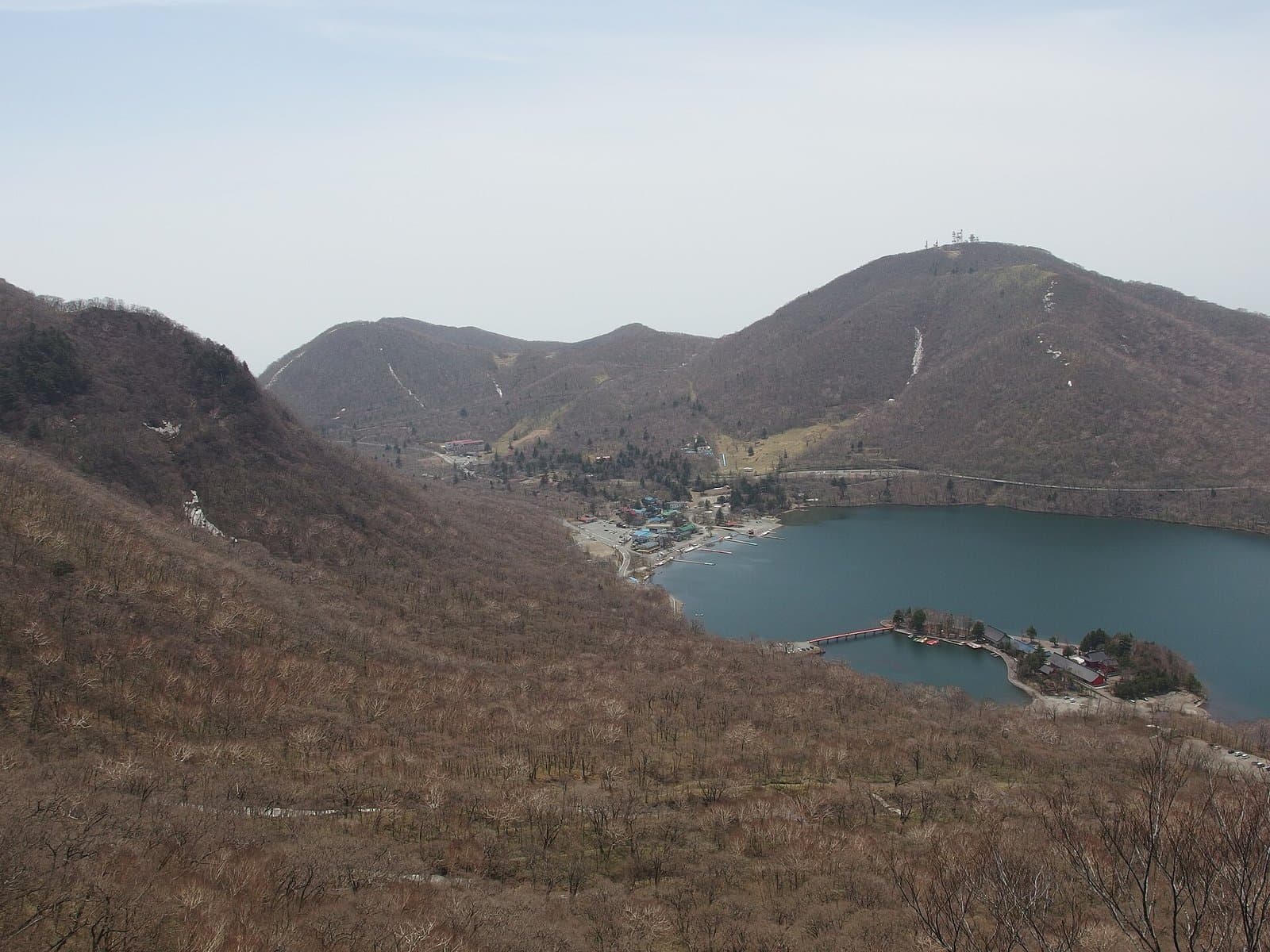 Lake Onuma seen from a mountain trail, with a red shrine on a small peninsula and bare early-spring trees
