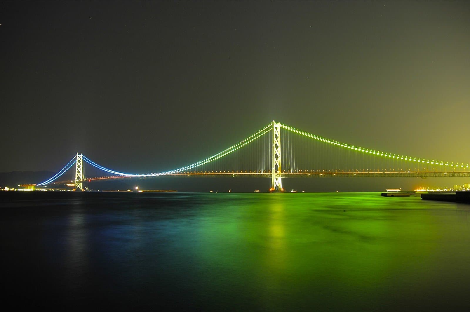 The long suspension bridge lit up in blue and green at night, reflected in the dark sea