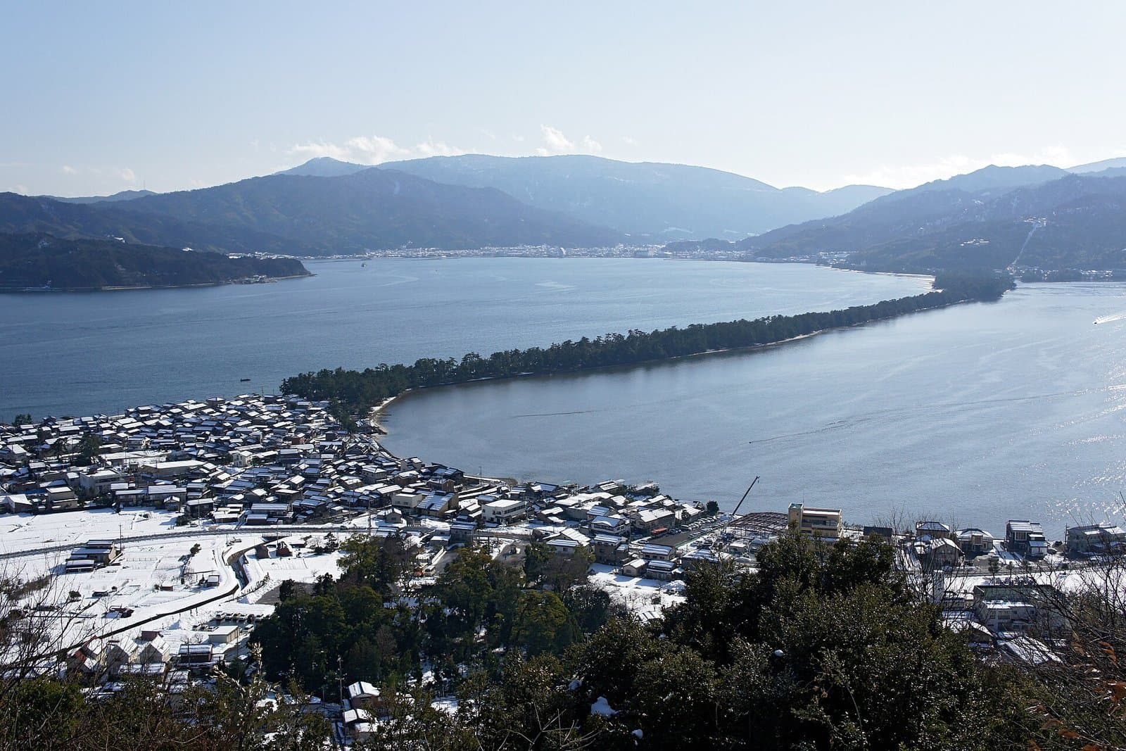 A long pine-covered sandbar stretching across a bay between mountains, seen from above a snowy town