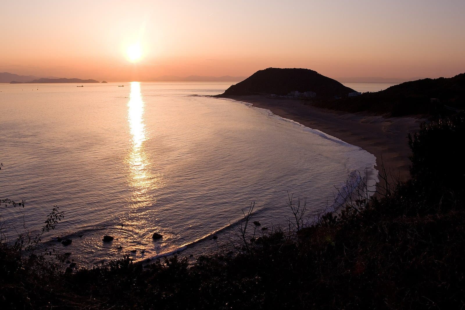 The sun setting over a calm sea beside a curving beach and a dark headland