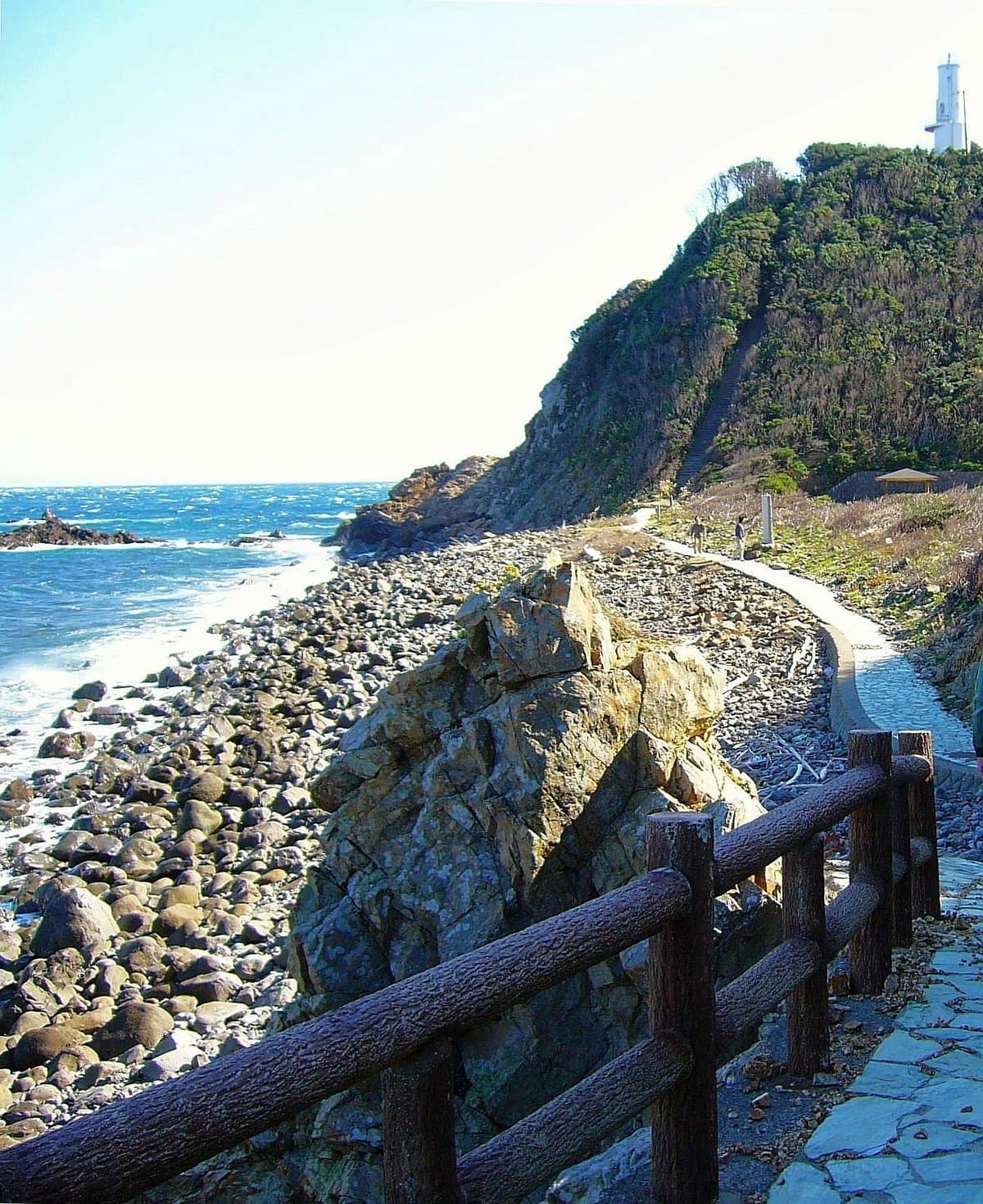 A path along a pebble beach toward a rocky cape with a white lighthouse