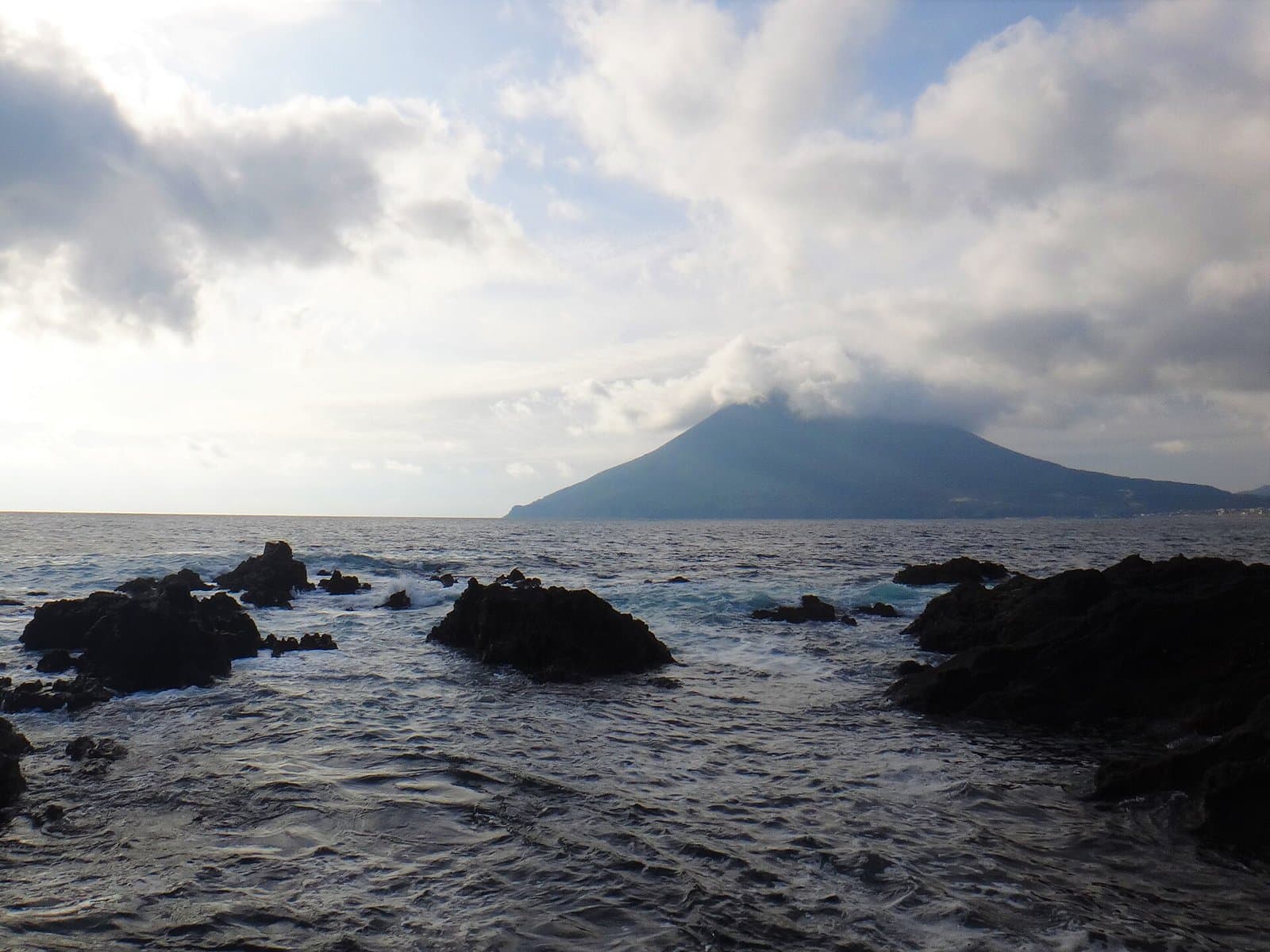 Dark rocks on the shore with a cone-shaped mountain across the sea