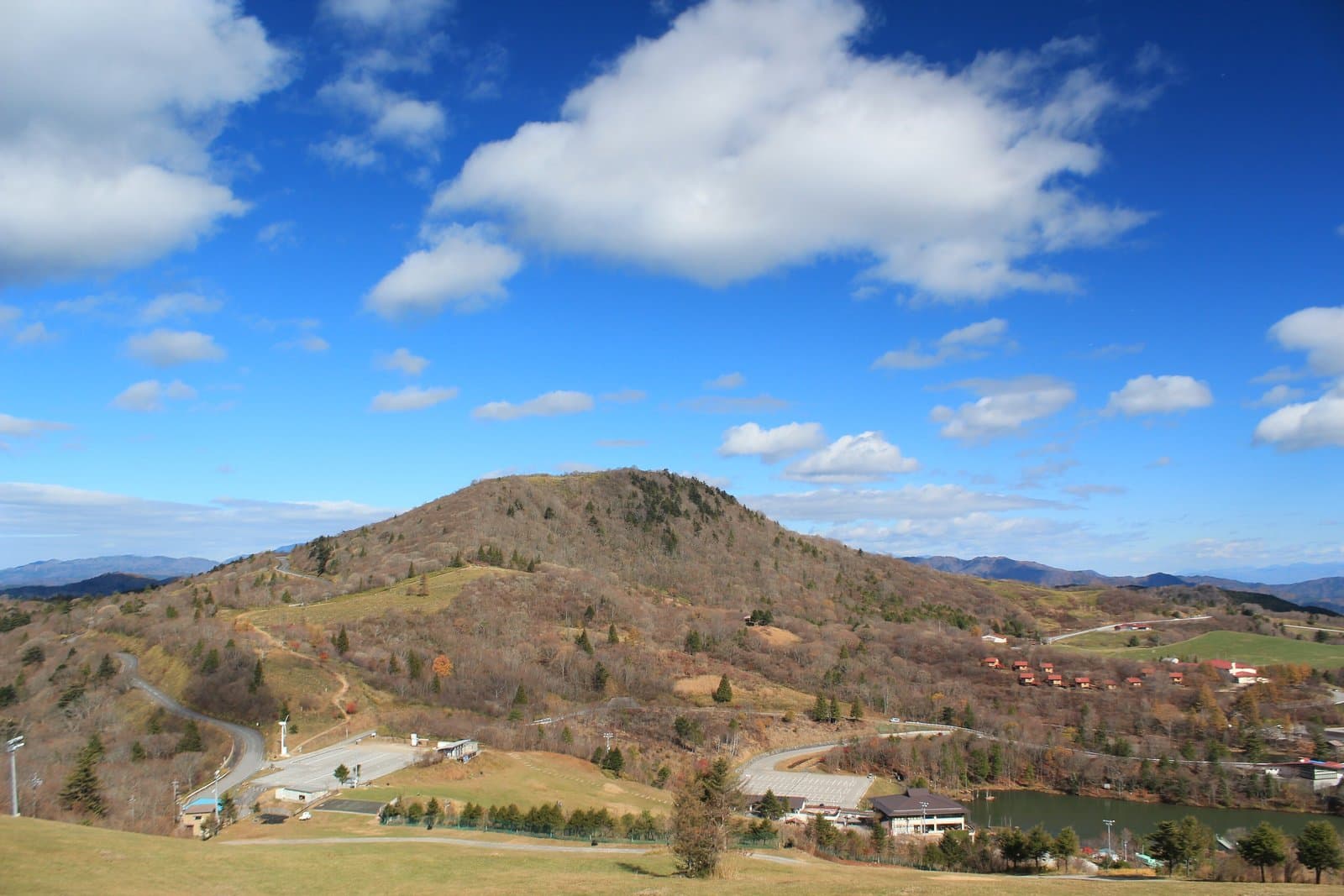A rounded mountain in late-autumn browns rising above a grassy highland with scattered trees and a small lake, under a blue sky with clouds