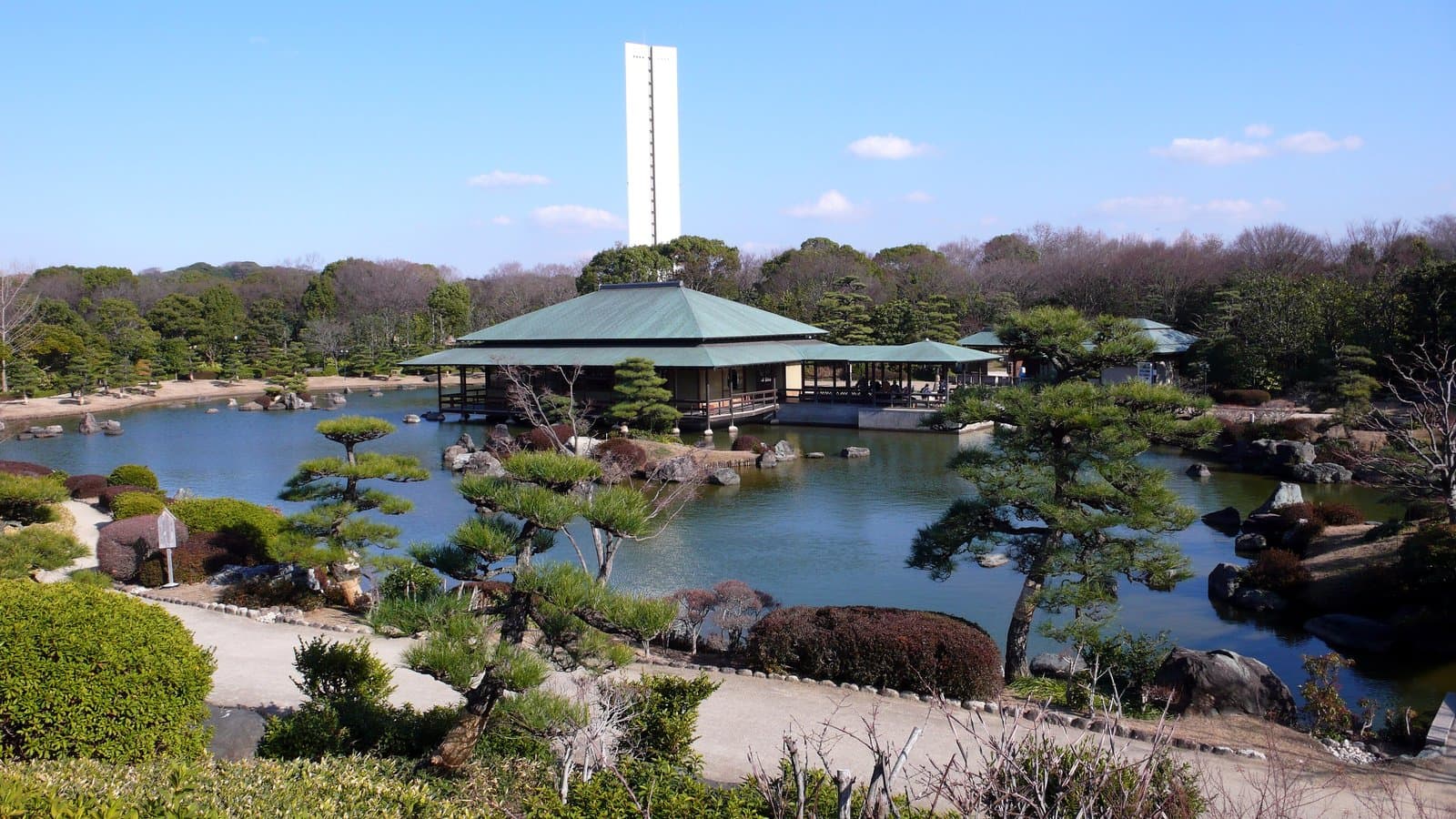 A Japanese garden pond with pruned pines and rocks, a pavilion on the water and a tall white tower beyond
