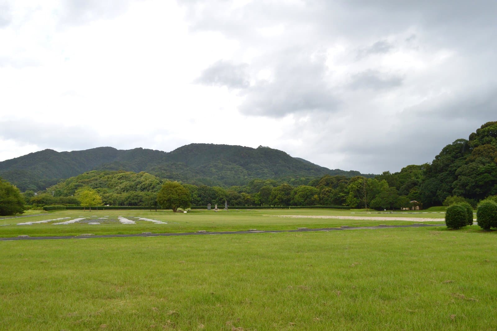 A wide grassy field with rows of foundation stones and stone monuments, below wooded hills