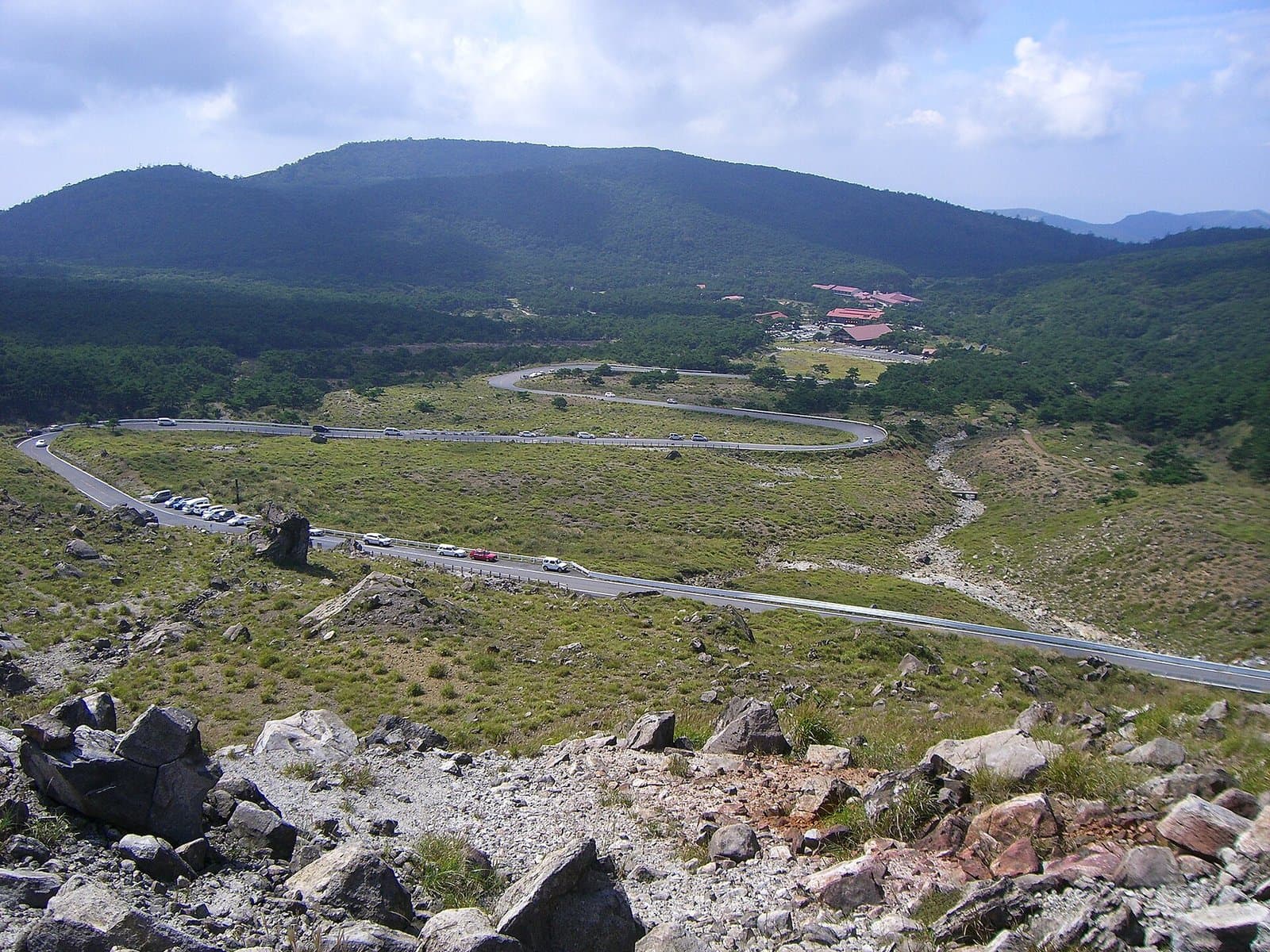 A winding road across a wide grassy plateau below a green mountain, seen from rocky high ground