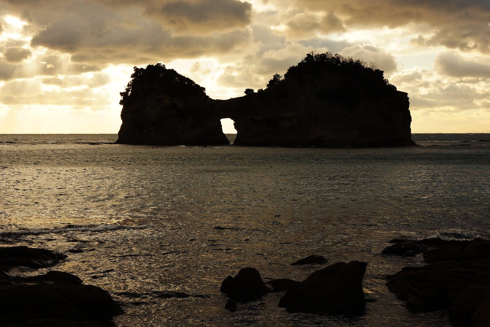 A rocky island with an arch-shaped hole, silhouetted against a cloudy sky over the sea