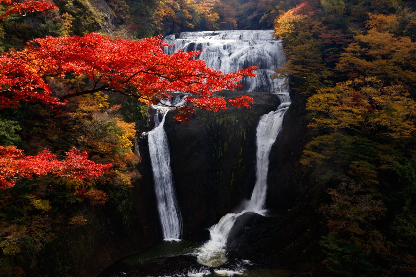A wide waterfall dropping down a dark rock face in tiers, framed by red and yellow autumn leaves