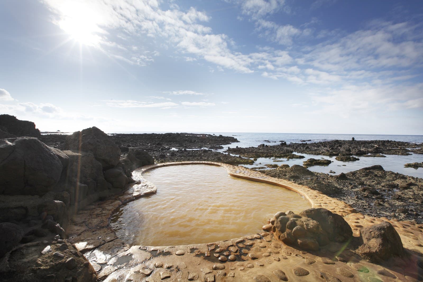 A gourd-shaped outdoor bath of brown water on the rocky seashore under a bright sky