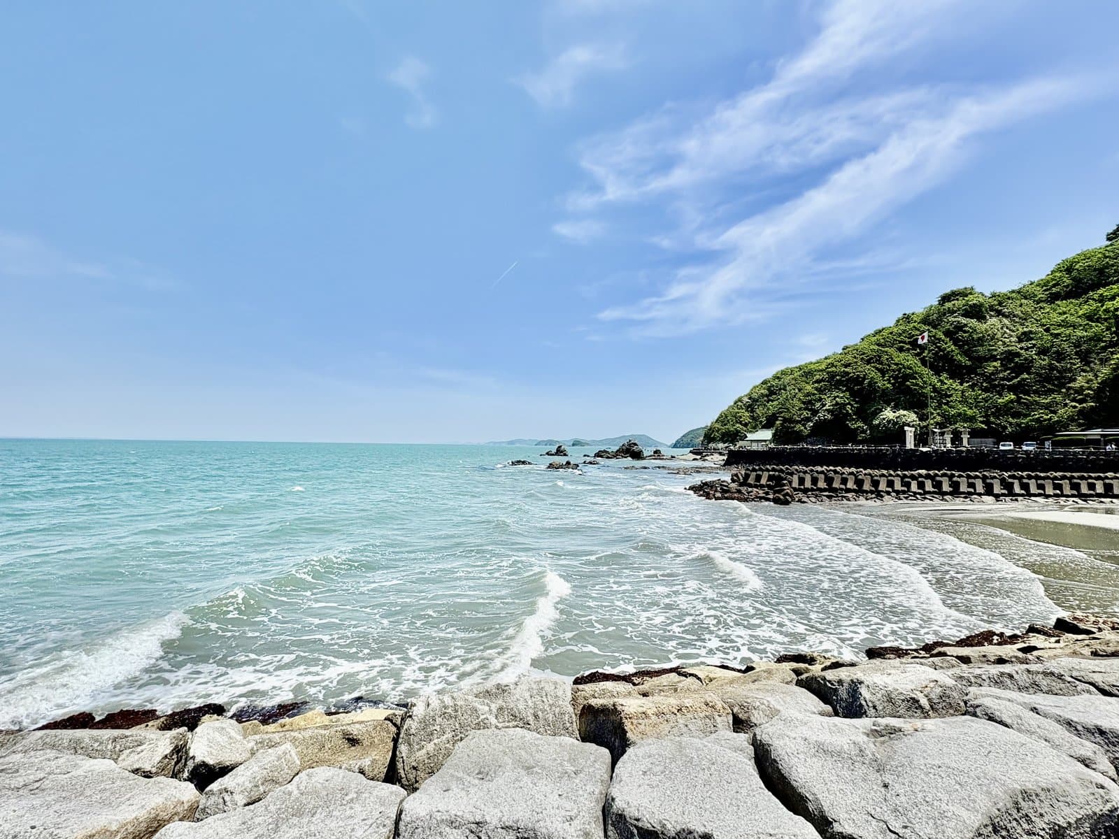 Waves rolling onto a beach below a green hill, seen from a rocky seawall