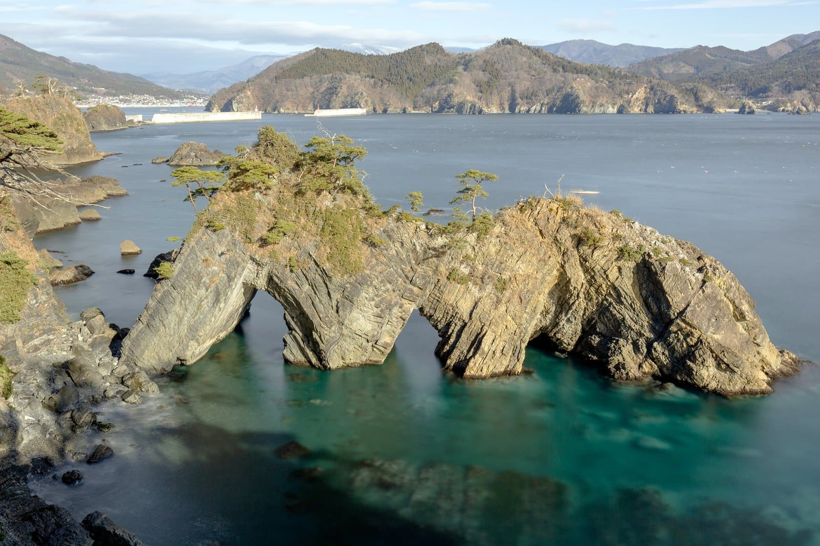 A rocky islet with three sea arches and pine trees on top, rising from clear turquoise water in a bay ringed by mountains