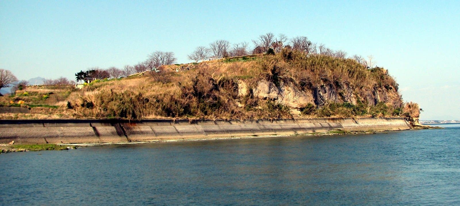 A low wooded headland with a stone seawall along a calm sea