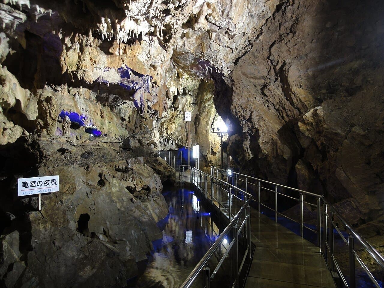 A lit walkway with railings winding through a limestone cave above still water reflecting blue lights