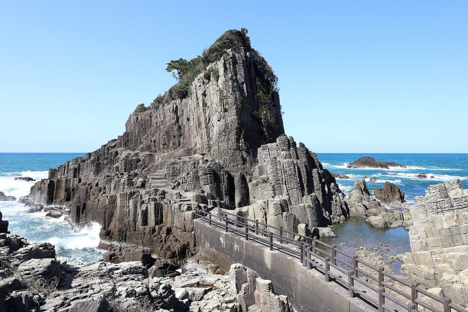 A tall rock of columnar basalt on a rocky shore, with a walkway with railings in front