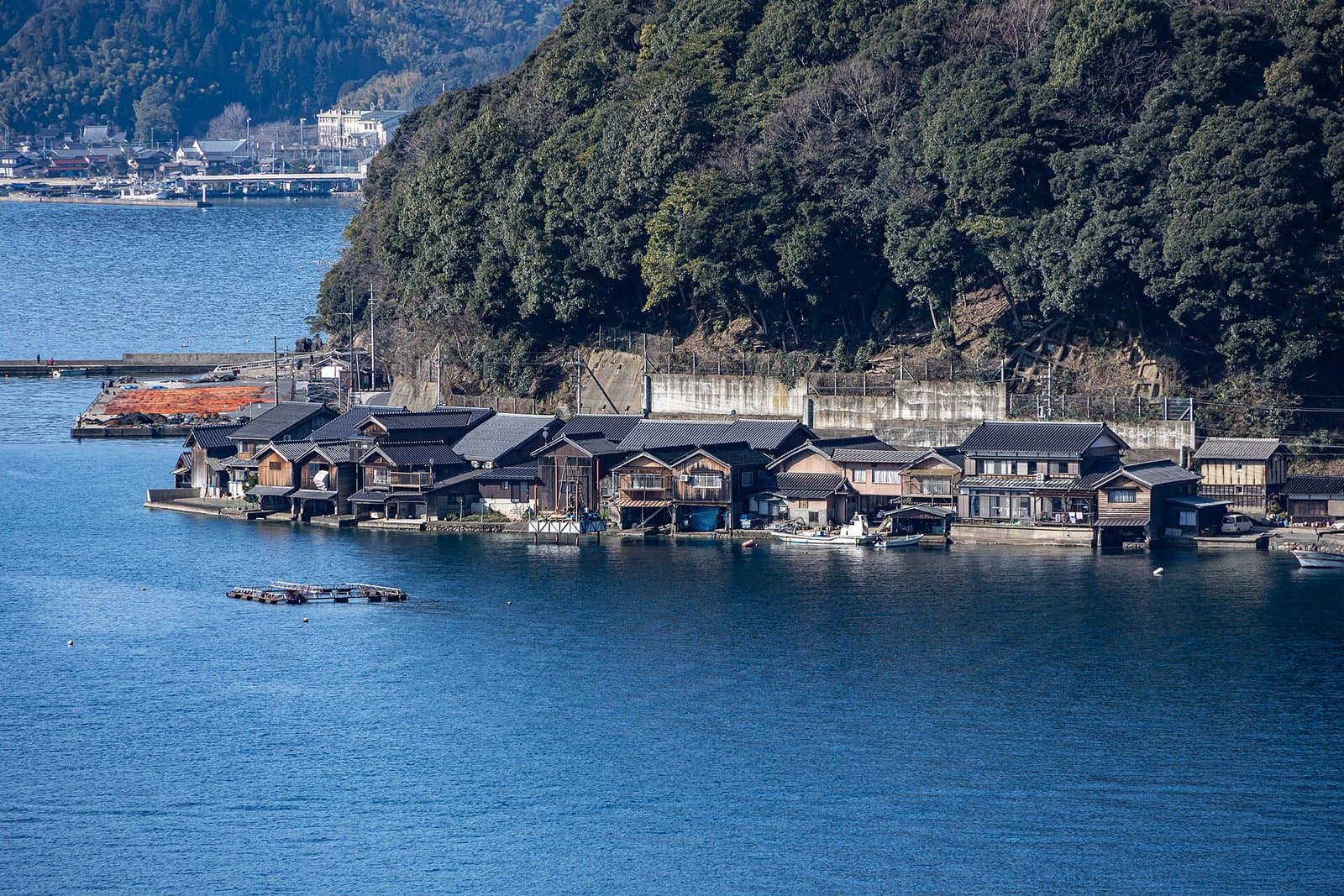 A row of wooden boathouses with tiled roofs along the water's edge below a wooded hill