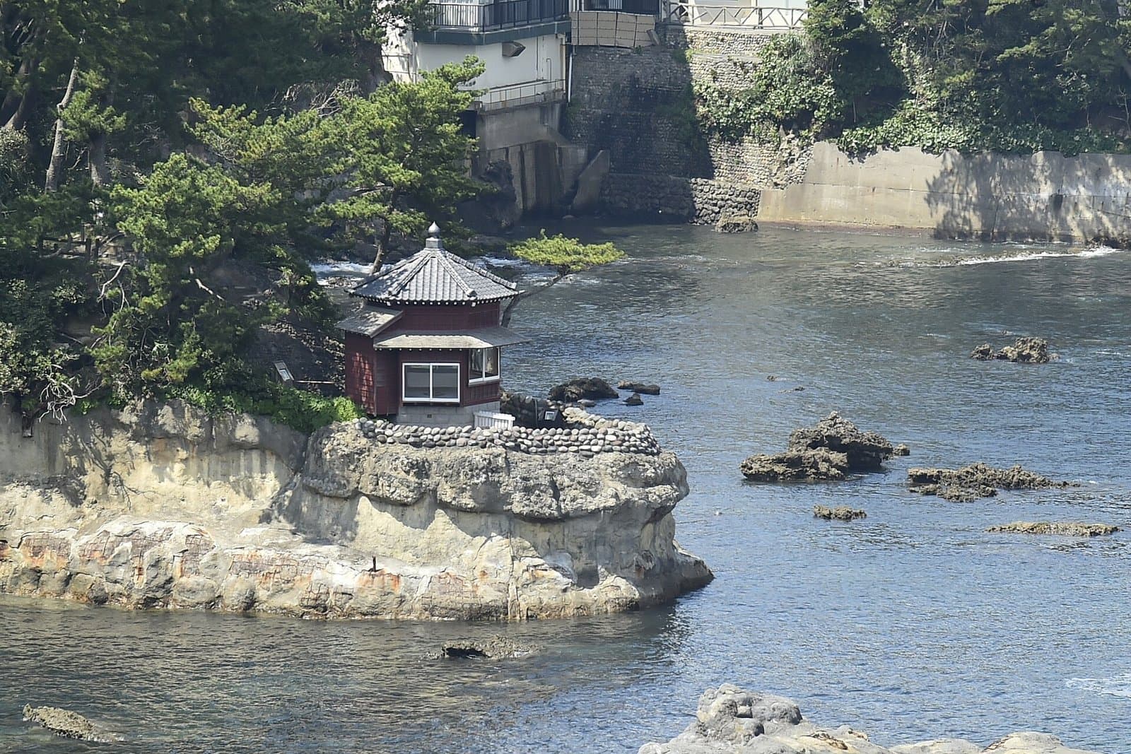 建在海上岩石上、有瓦屋頂的紅色六角形小堂與後方的松樹
