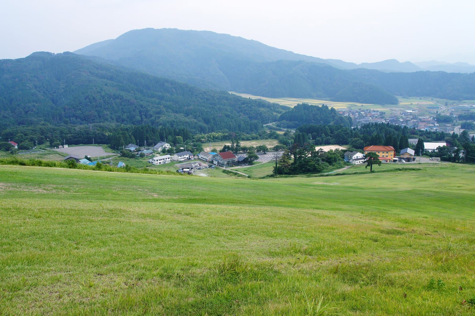 A green grassy slope above a village of lodges, with rice fields and mountains beyond