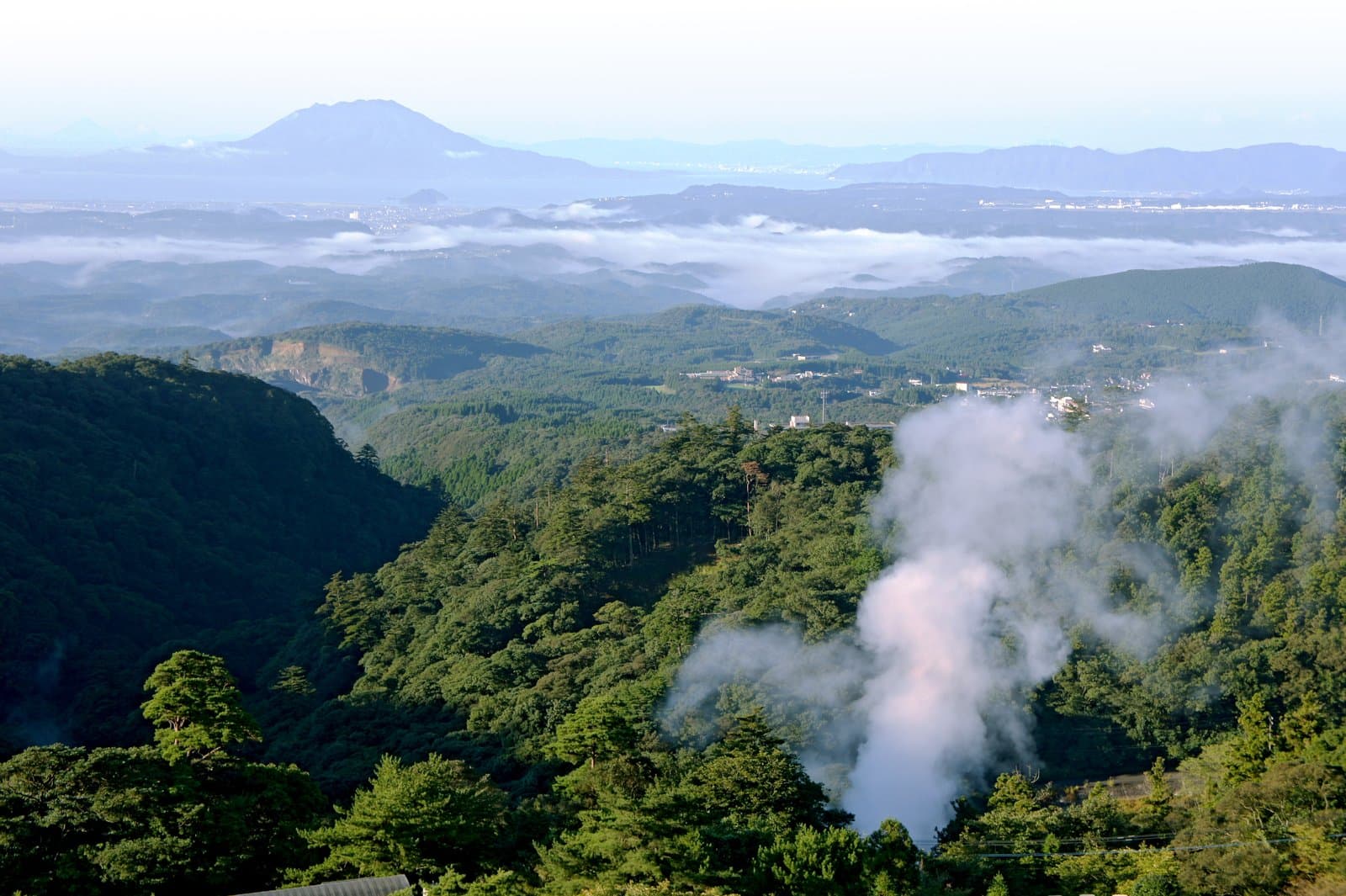 Steam rising from a forested slope, with hills, a sea of clouds and Sakurajima in the distance