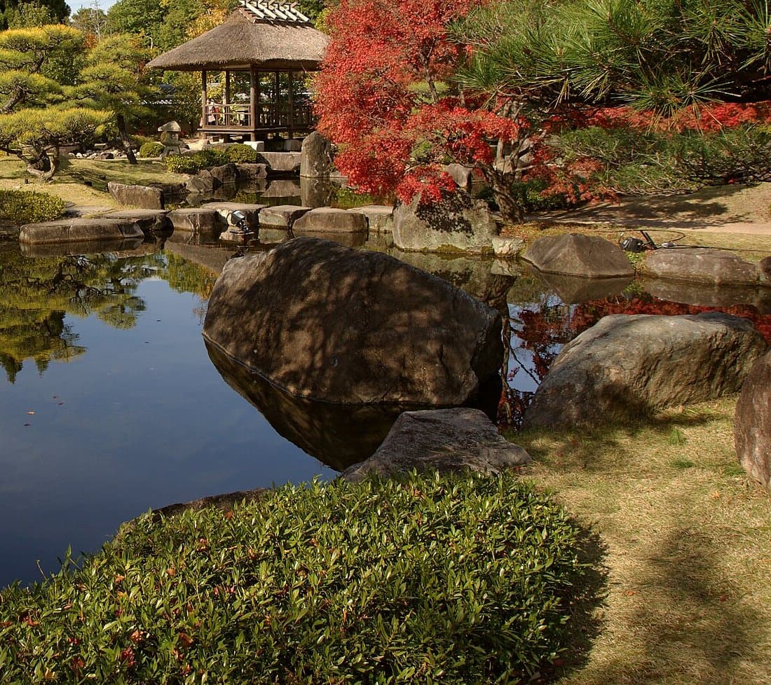 Red maple leaves over a pond with large rocks, with a thatched arbour behind