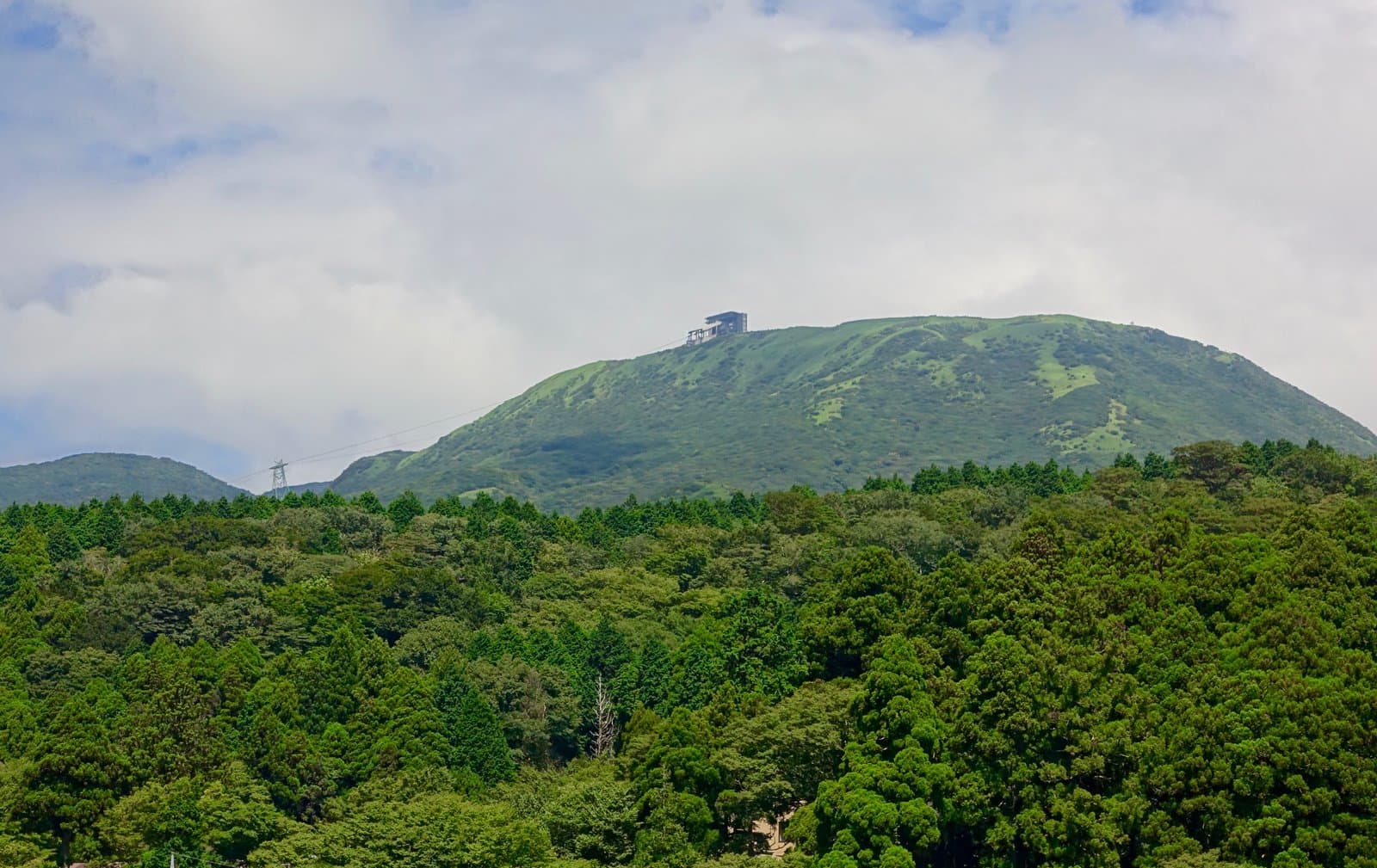 A grassy mountain top with a small ropeway station, above a dense green forest