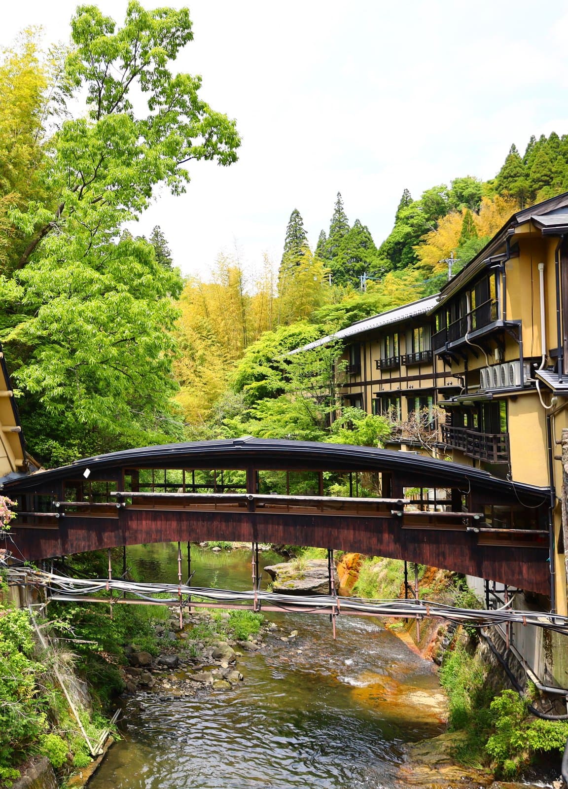 A covered wooden bridge over a rocky stream between green trees and a timber-framed inn