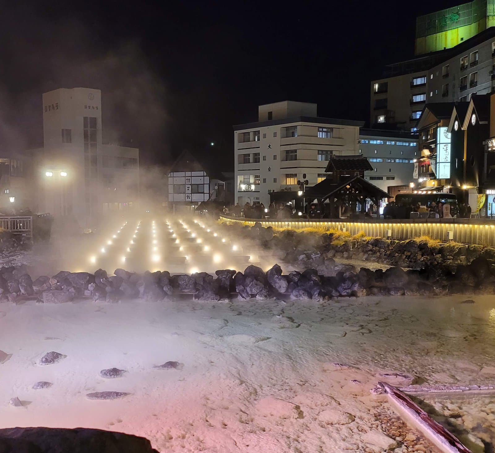 Steam rising from the lit-up hot-spring channels of Yubatake at night, with inns behind