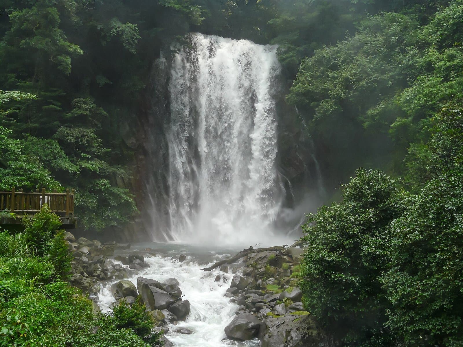 A white waterfall pouring into rocks in a green forest, with a wooden viewing deck on the left