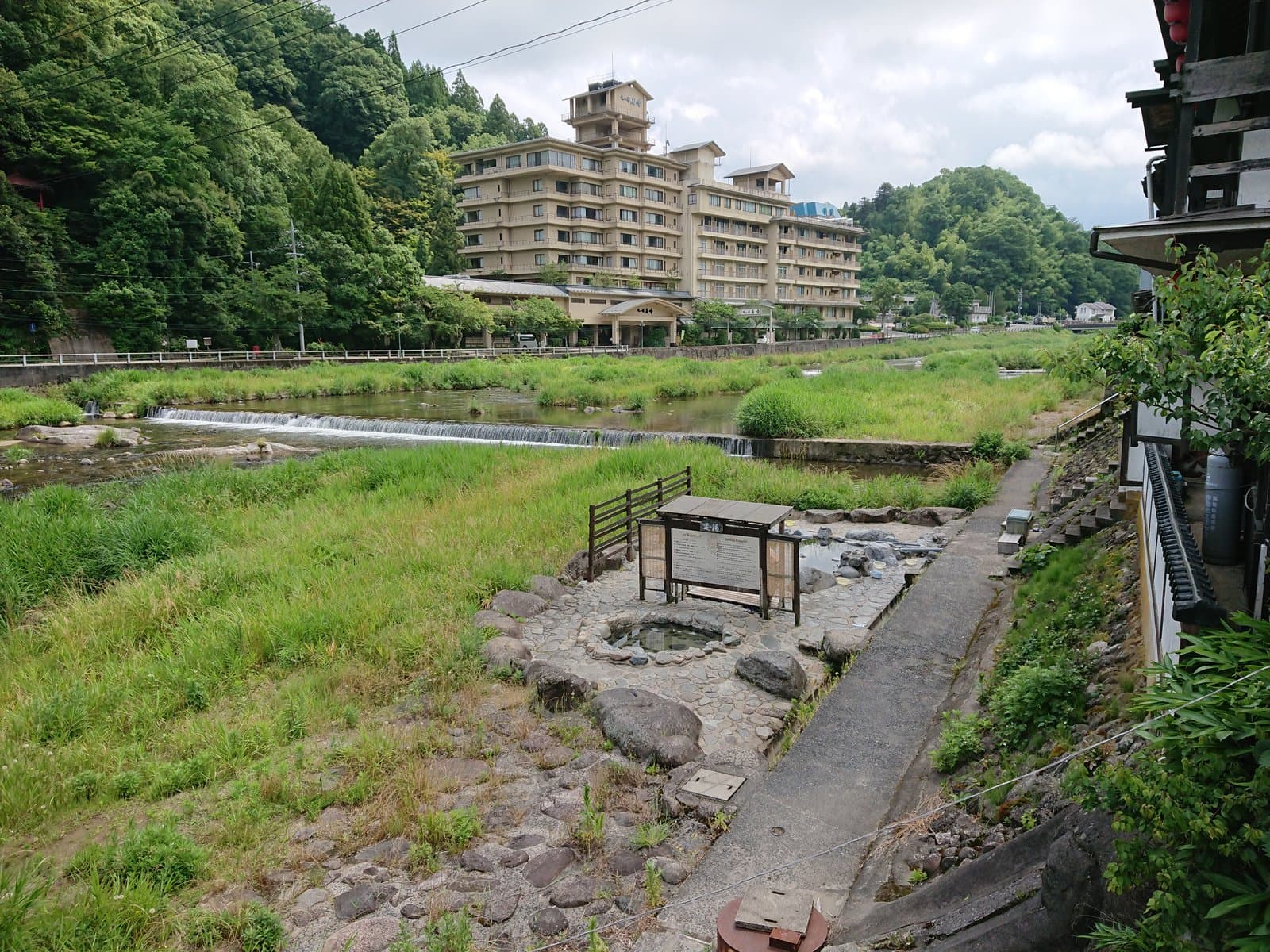 A stone open-air bath by a grassy riverside, with a weir and a hotel beyond