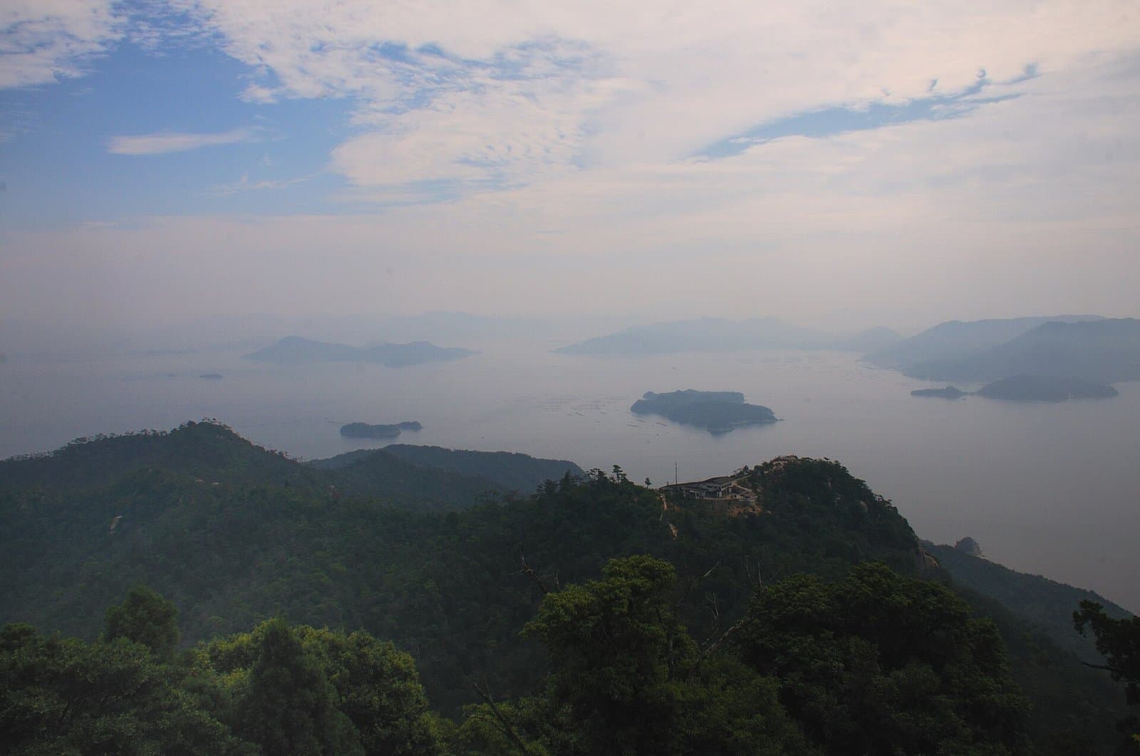 Forested ridges in the foreground and islands in a hazy Seto Inland Sea under a cloudy sky