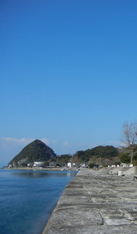 An old stone wharf along a calm blue sea, with a pointed wooded hill beyond