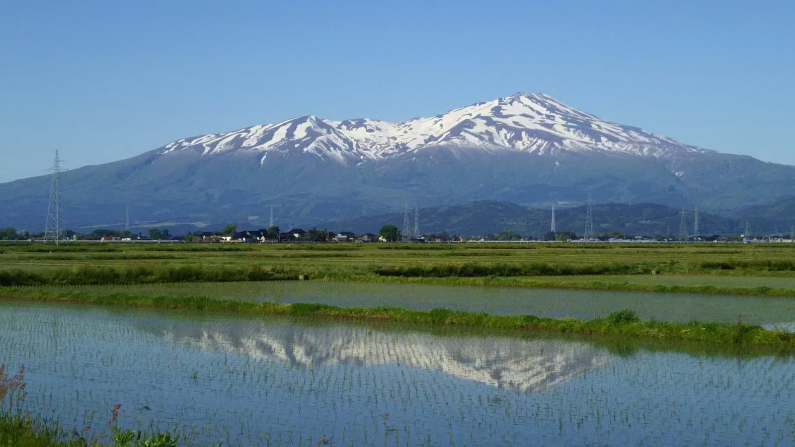 A snow-streaked mountain reflected in flooded rice paddies, with a row of houses at its foot