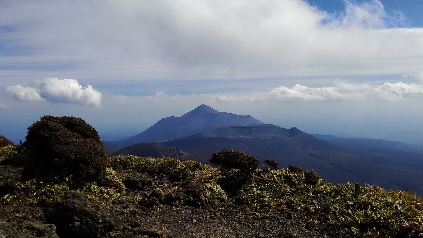 A dark volcanic peak under a cloudy sky, seen across shrubs from a rocky mountain trail