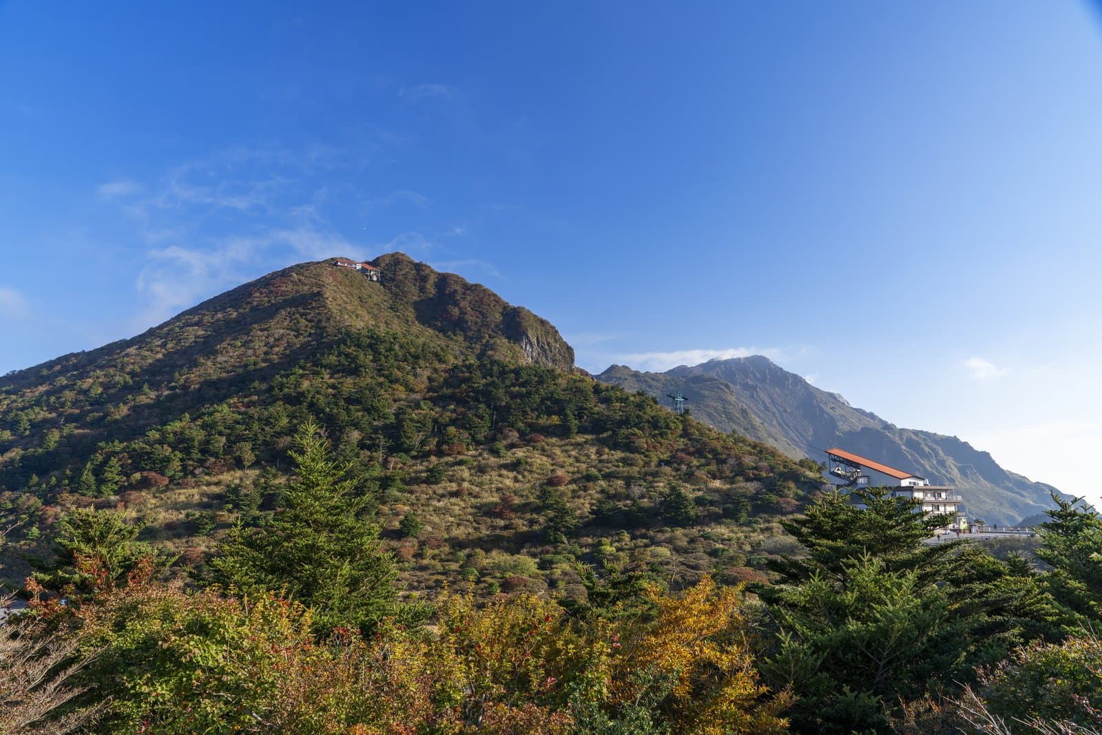 A wooded mountain in early autumn colours under a blue sky, with a ropeway station on its ridge