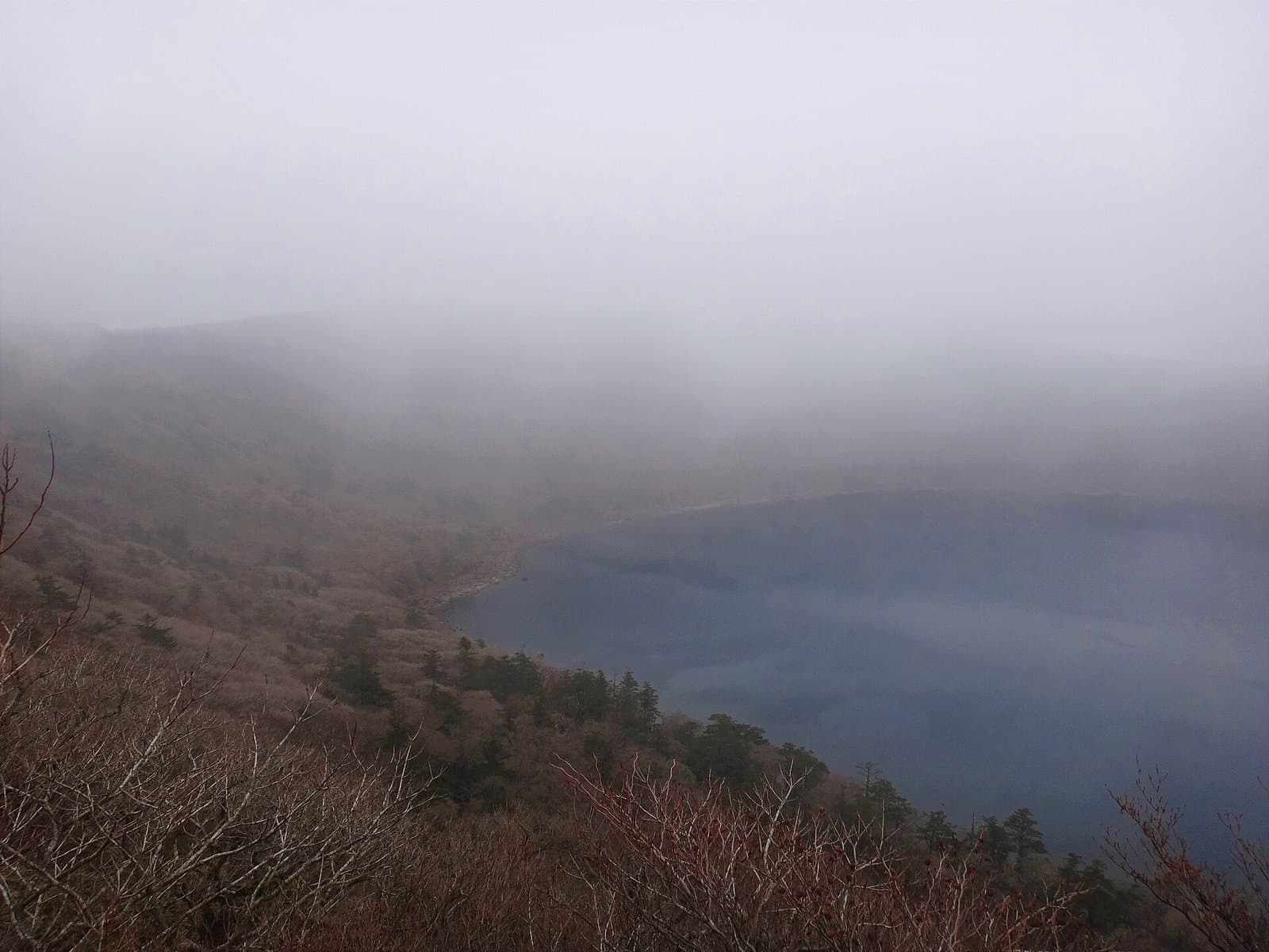 A crater lake half hidden in fog, seen from a slope with bare shrubs