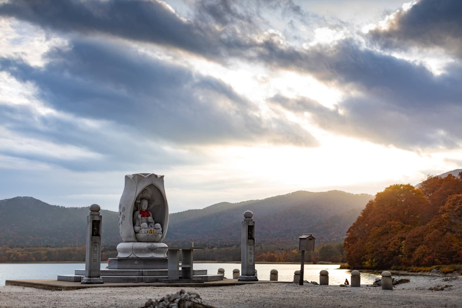A stone Jizo statue on the lakeshore at dusk, with autumn-coloured hills