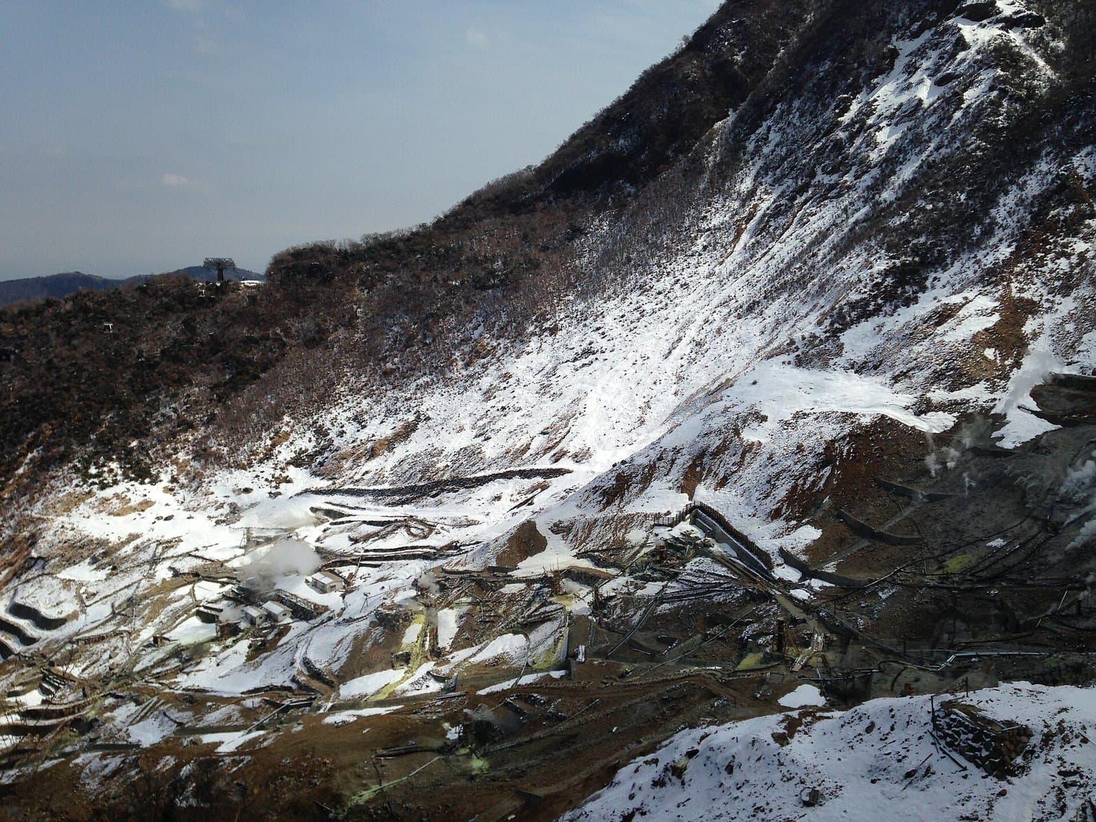尾根にロープウェイの鉄塔が立つ雪の急斜面と、その下で噴気が上がる硫黄色の大涌谷
