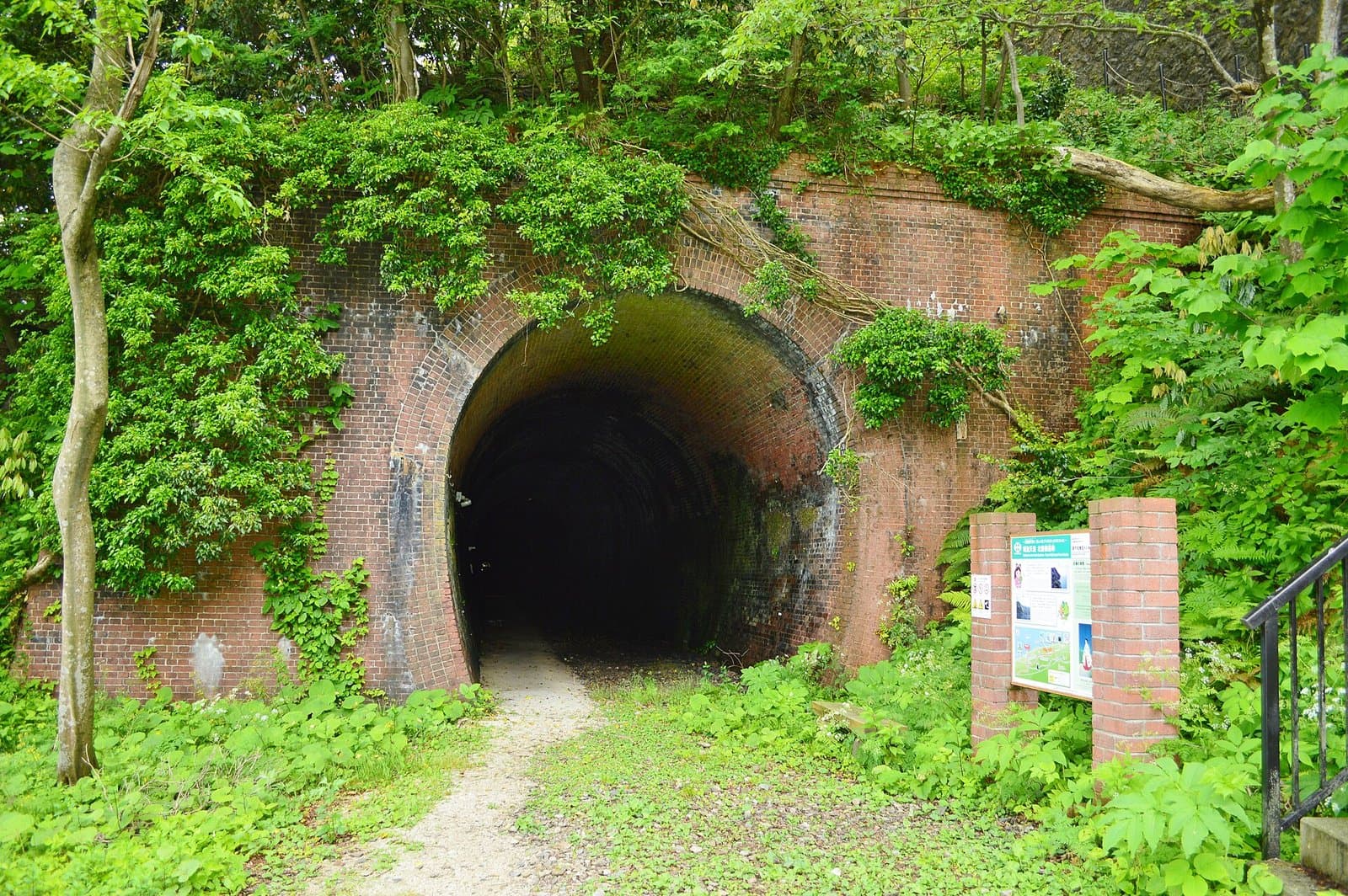 The arched entrance of an old red-brick tunnel overgrown with plants, with an information board