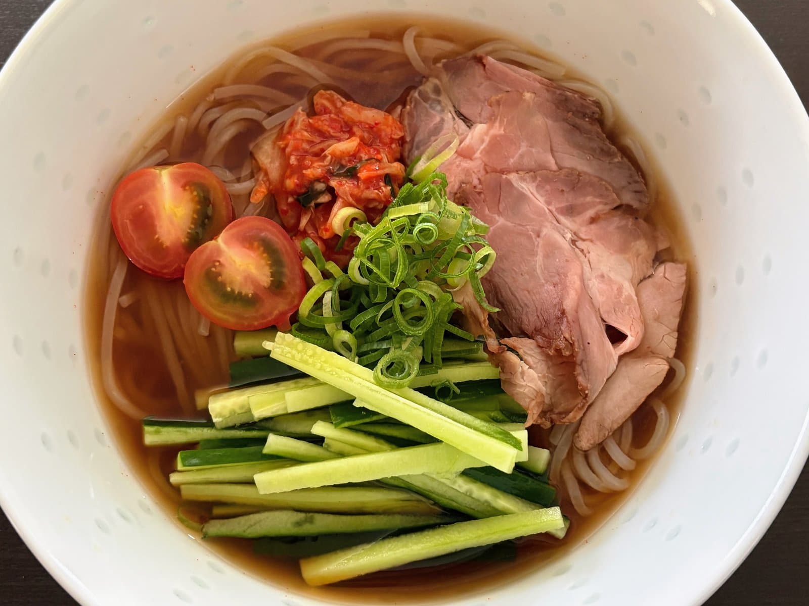 Cold noodles in clear broth topped with sliced beef, kimchi, cucumber, tomato and green onion