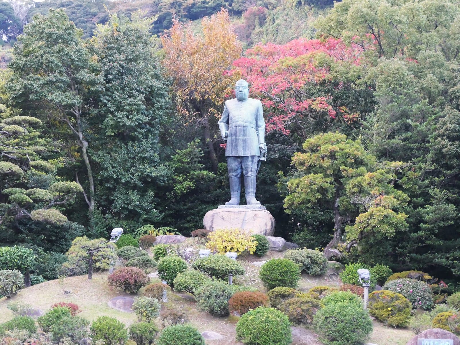 刈り込まれた低木の庭の上、石の台座に立つ軍服姿の大きな銅像と、後ろの色づいた木々