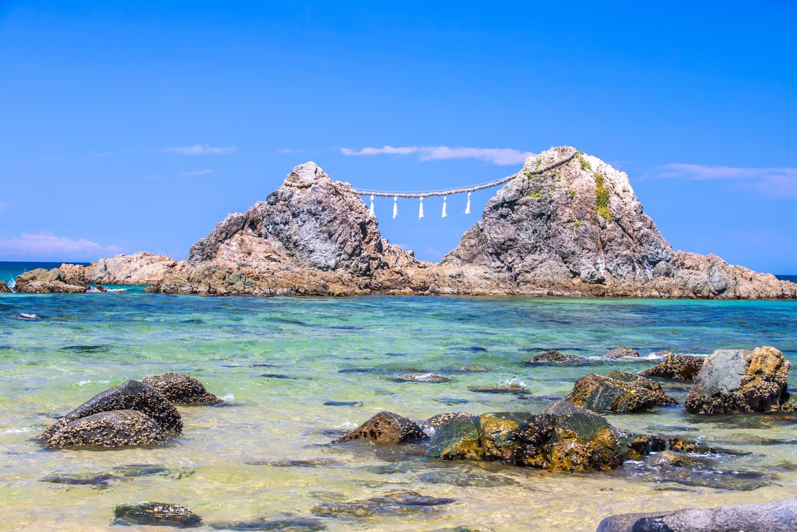 Two rocky islets joined by a thick sacred rope with white paper strips, in clear shallow sea