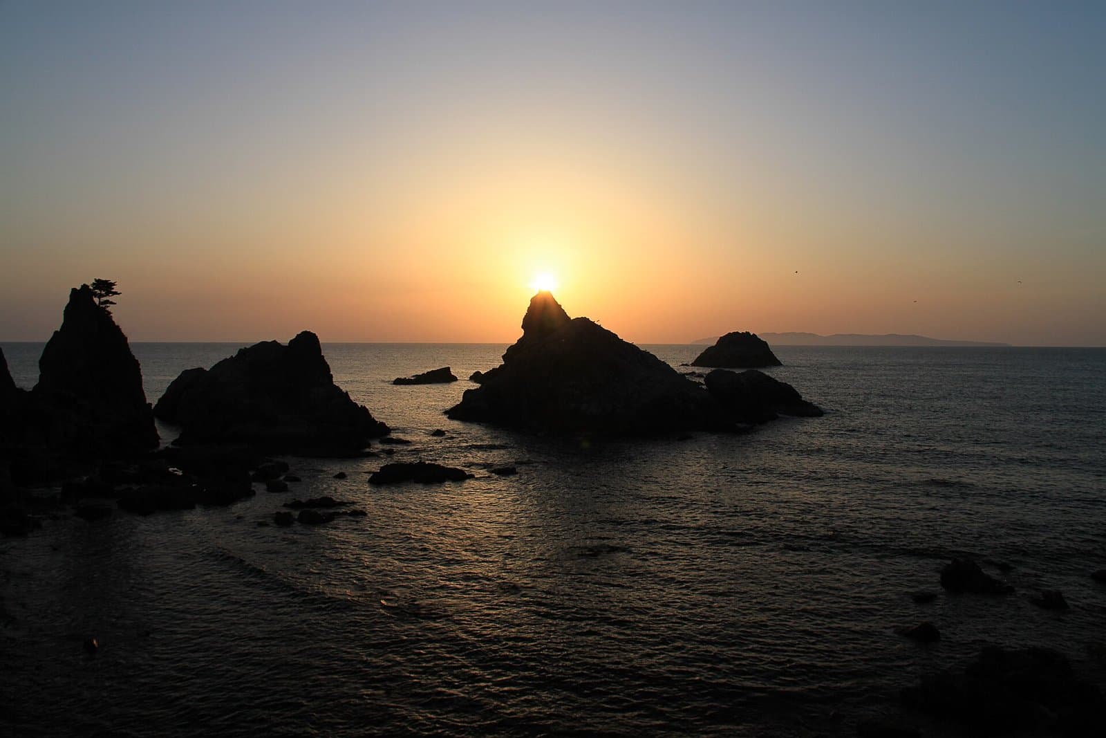 The sun setting behind dark rocks standing in the sea