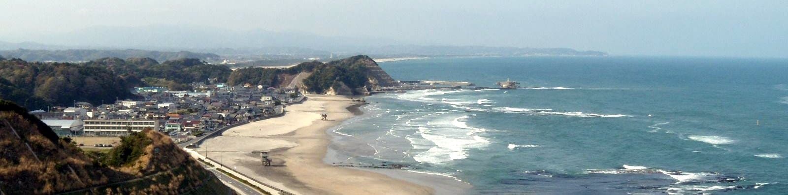 A curving sandy beach and a small town along a blue sea with white surf, seen from high above