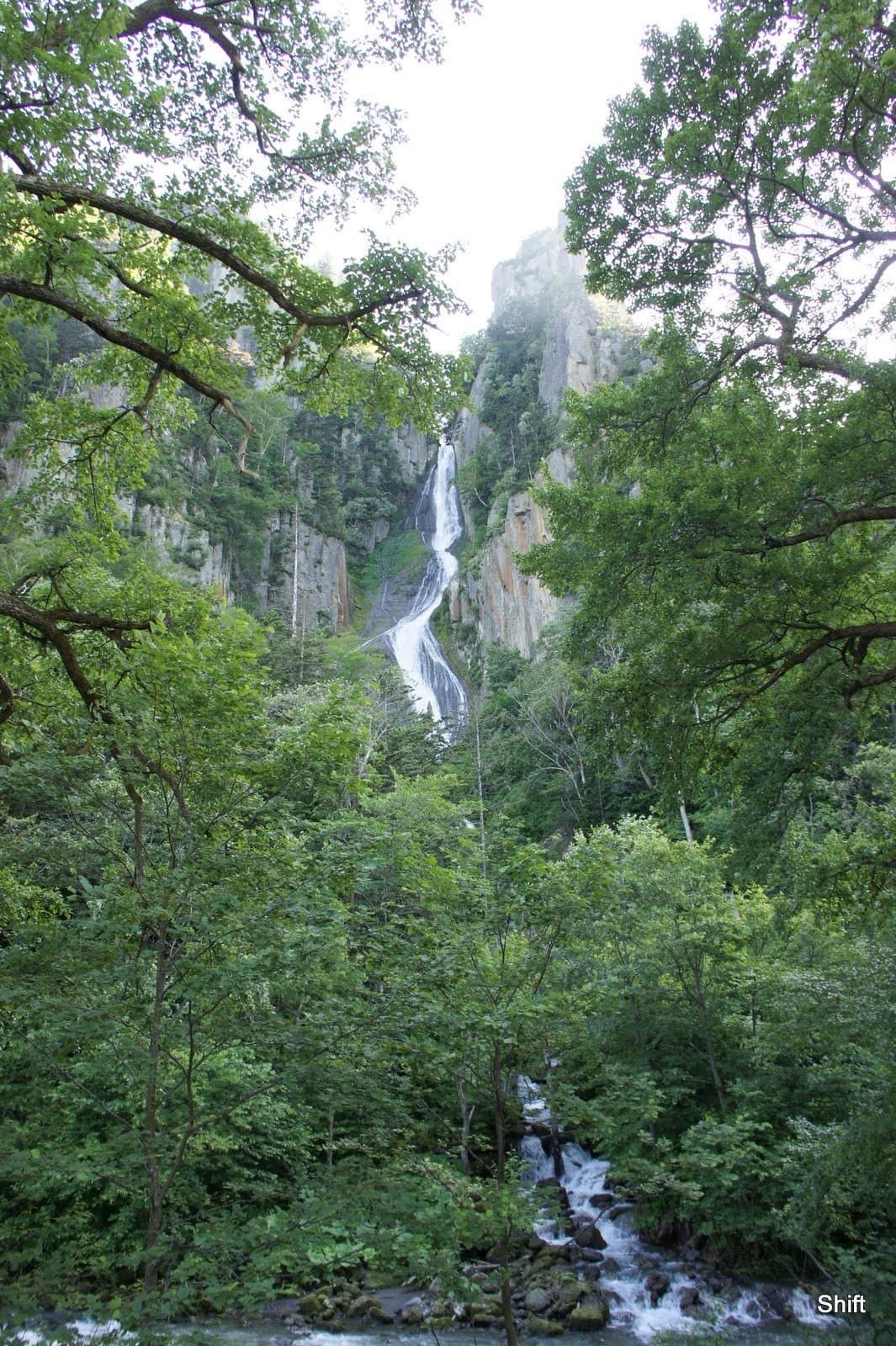 A slender waterfall falling down a cliff above green forest and a stream