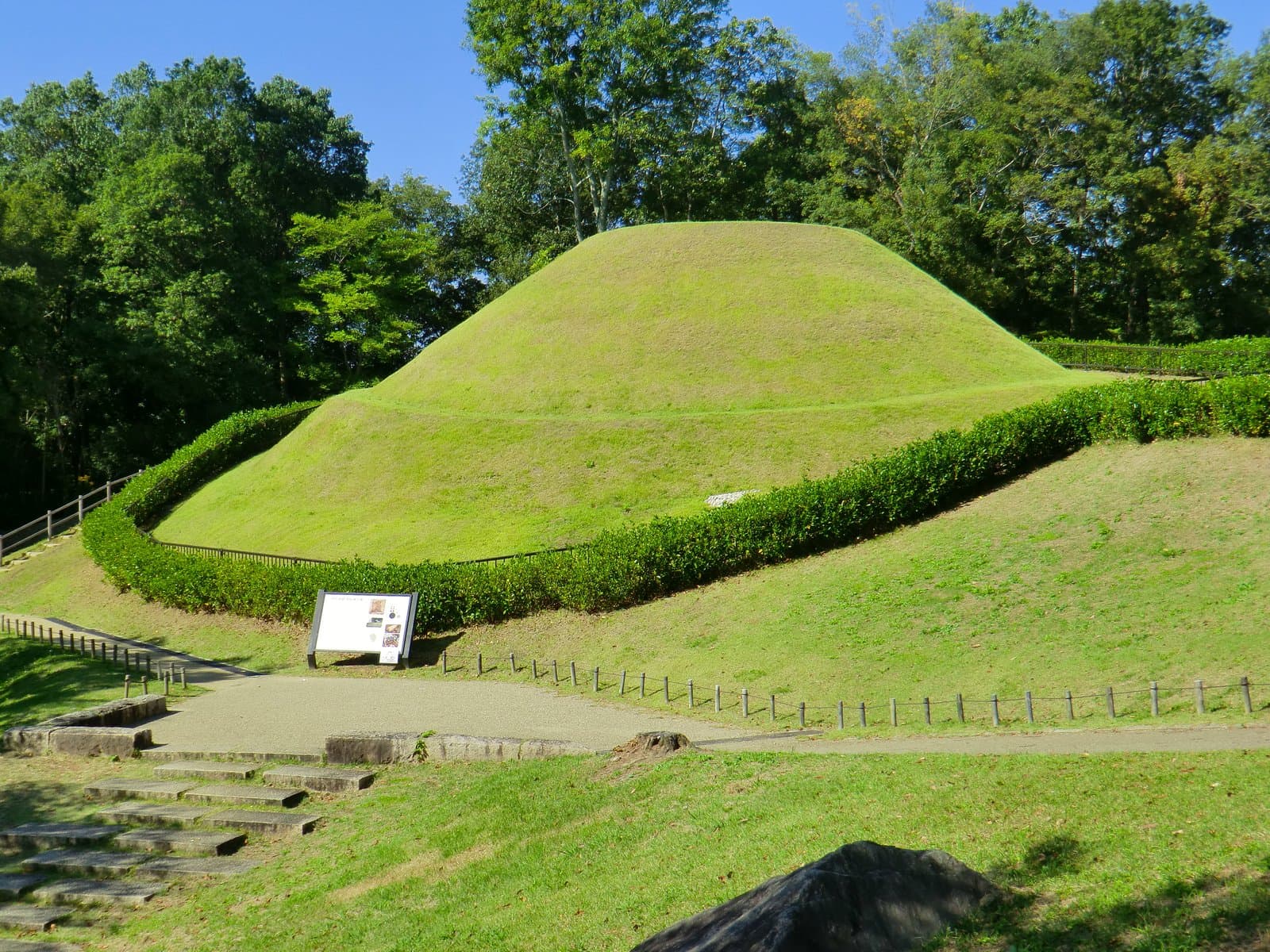 A round grassy burial mound in two tiers, ringed by a clipped hedge and backed by trees