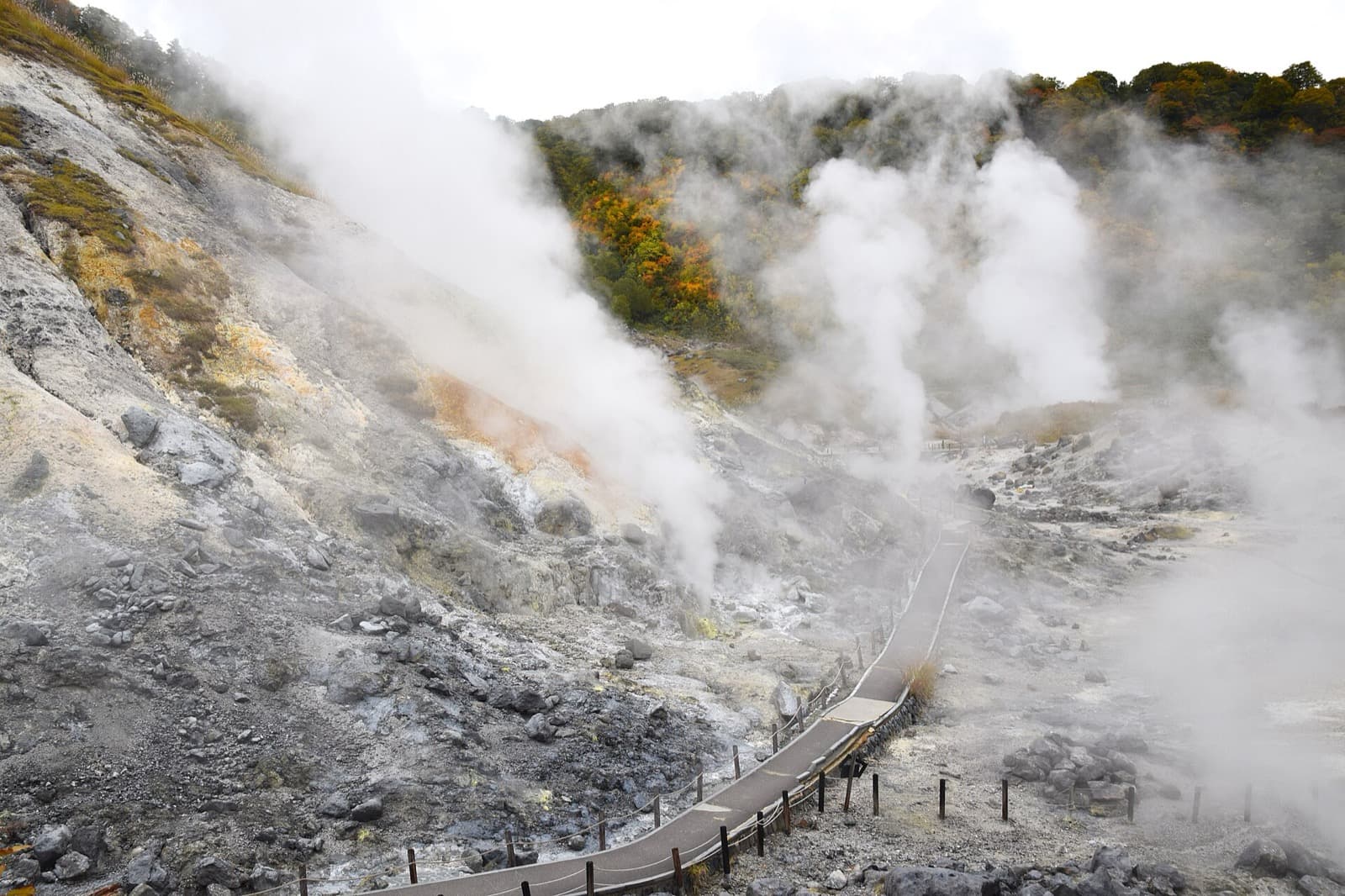 Steam rising from a rocky volcanic slope, with a paved walkway and autumn trees