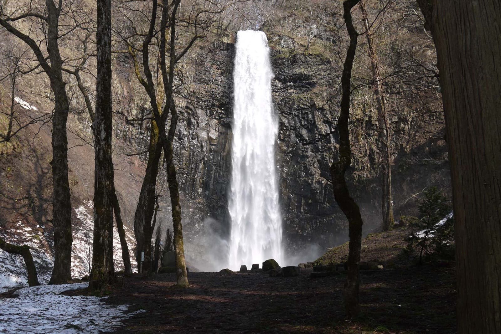 A tall waterfall dropping down a dark rock face, seen between trees with patches of snow