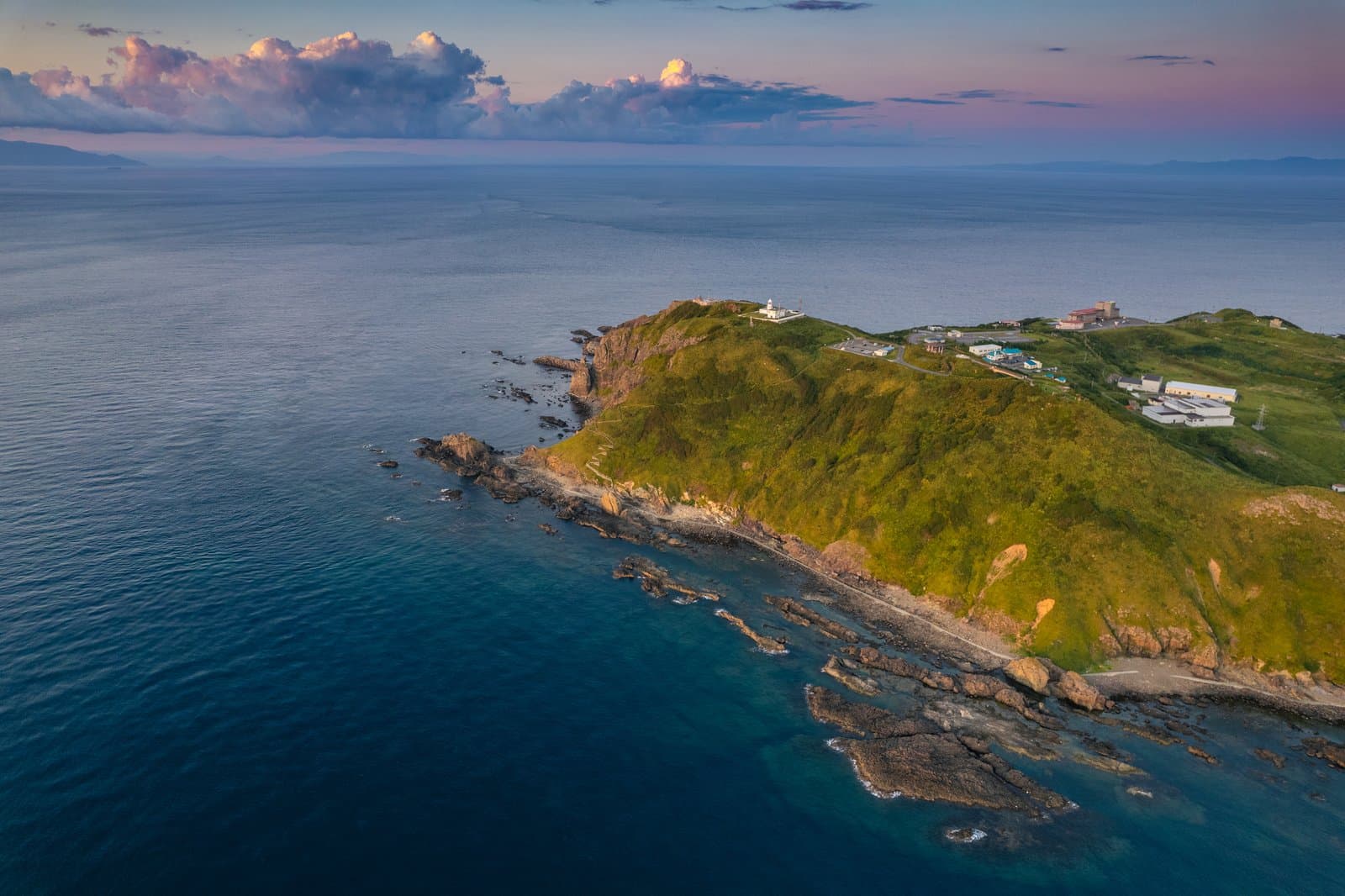 A green cape with a white lighthouse on a calm sea at dusk, seen from above