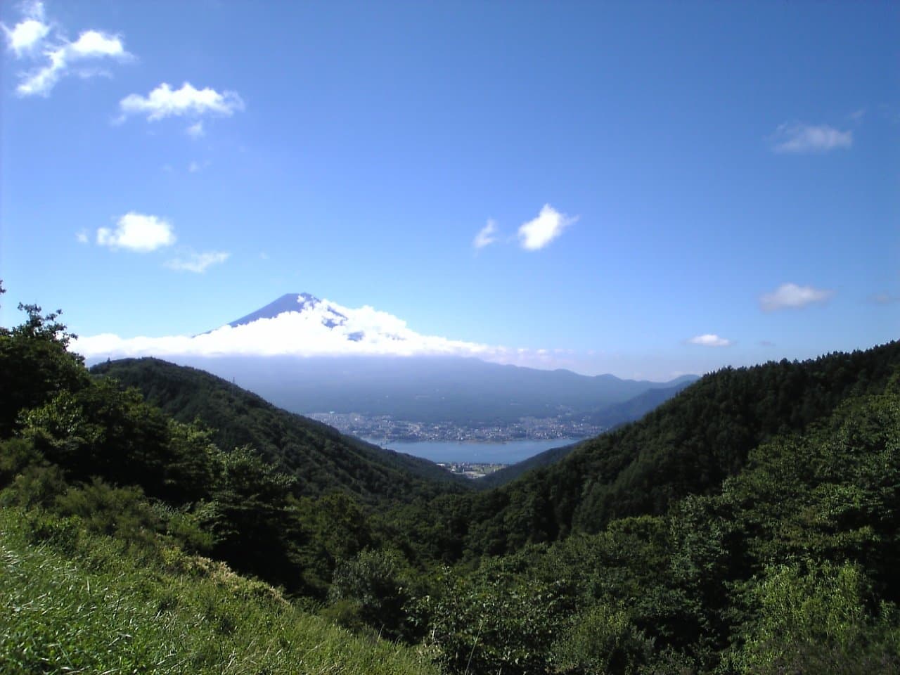 Mt. Fuji rising above clouds beyond a green valley and a glimpse of a lake, under a blue sky