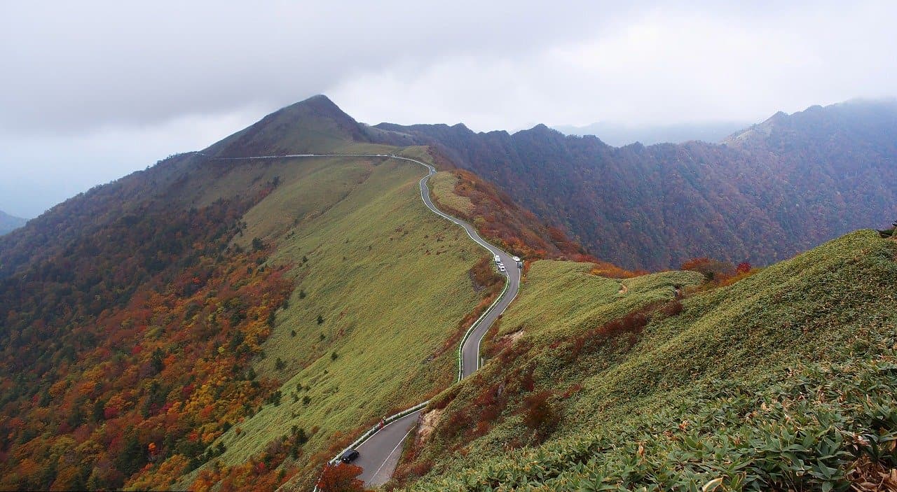 A narrow road winding along a mountain ridge with autumn-coloured slopes