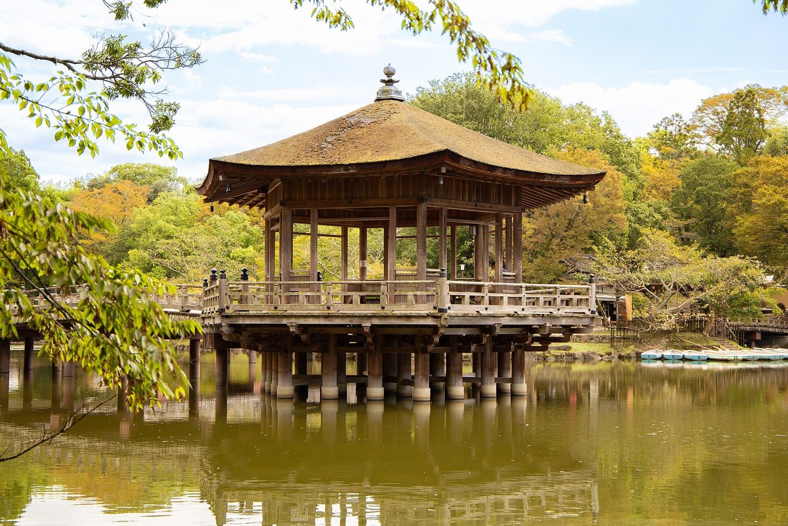 A hexagonal wooden pavilion with a bark roof standing in a pond, surrounded by trees